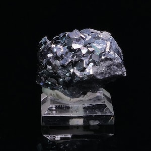 Può includere: Un gruppo di cristalli di ematite neri iridescenti su un supporto in vetro trasparente. I cristalli hanno un'opalescenza metallica e sono disposti in modo casuale.