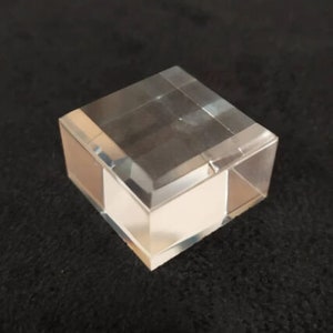 Puede incluir: Un expositor de cristal transparente con base cuadrada y parte superior rectangular. El expositor está hecho de cristal grueso y transparente y tiene un acabado pulido.