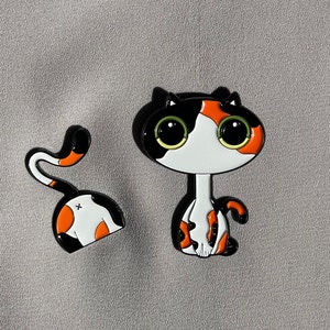 May include: Two black and orange enamel pin badges of a cat with large eyes. One cat is sitting upright with its tail curled around its legs. The other cat is curled up with its tail wrapped around its body.
