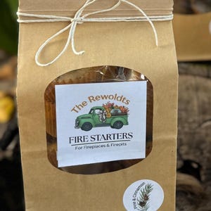 May include: A brown paper bag tied with twine, with a clear window showing fire starters. The label says "The Rewoldts FIRE STARTERS" with a green truck graphic. A round sticker with pine and cinnamon is on the bag.