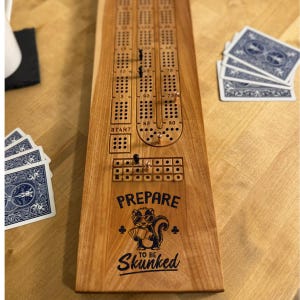 Può includere: Tavola da cribbage in legno con grafica di puzzola e la scritta "Prepare to be Skunked". La tavola presenta piste di punteggio, fori per pioli e pioli. Carte da gioco visibili. Un classico gioco per due o più giocatori.