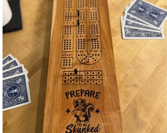 May include: A wooden cribbage board with a skunk graphic and the text "Prepare to be Skunked." The board has scoring tracks, peg holes, and pegs. Playing cards are visible. A classic game for two or more players, ideal for family game nights.