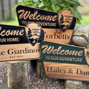 Puede incluir: Carteles de bienvenida de madera con texto tallado y emblemas del Servicio de Parques Nacionales. Un letrero dice "Bienvenido a nuestro hogar" con "The Giardinos" y "EST 2005". Otro dice "Bienvenido a nuestra aventura" con "Hailey & Daniel" y "EST 2020".