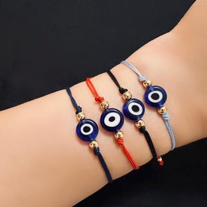 Puede incluir: Cuatro pulseras con cuentas azules de ojo turco. Cada pulsera presenta un cordón de diferente color: azul marino, rojo, negro y gris claro. Las cuentas de ojo turco son azules con un centro blanco y detalles dorados.