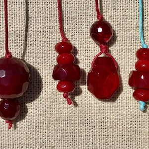 May include: Four red gemstone pendants on red and blue cords. The pendants have various shapes, including faceted spheres and geometric forms. The gemstones are strung on thin cords, creating a simple, elegant design. The background is a textured, neutral fabric.