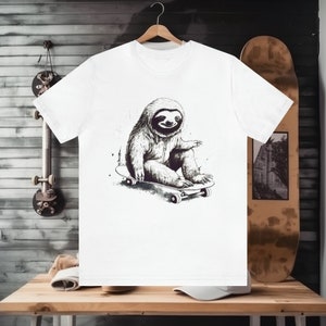 May include: A white t-shirt with a black and white illustration of a sloth riding a skateboard. The sloth is sitting on the skateboard with its arms outstretched and its legs bent. The skateboard has black wheels and a black deck.