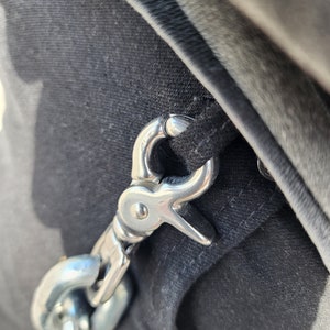 Puede incluir: Un gancho y una cadena de metal plateado unidos a una correa de tela negra.