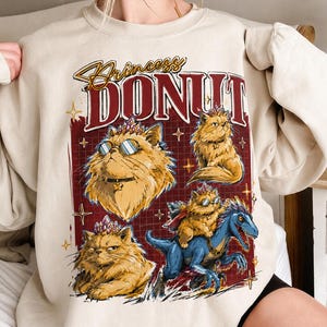 May include: A beige crewneck sweatshirt featuring a graphic print with the text Princess DONUT. The design includes three illustrations of a fluffy orange cat wearing a crystal crown and a cat riding a blue dinosaur, set against a dark red grid.