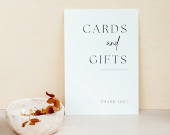 May include: A white sign with the words "CARDS and GIFTS" in black script and a line underneath. Below the text is "THANK YOU!". A small, textured white bowl with dried leaves sits to the left.