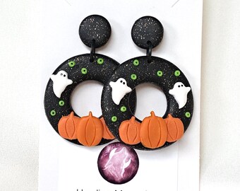 May include: A pair of black circular earrings with white speckles, featuring small white ghost figures, orange pumpkins, and tiny green dots. The earrings are displayed on a white card with the text Howling Moon Clay and an email address.