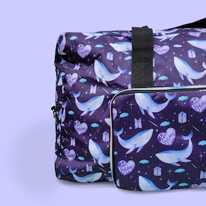 May include: A purple travel bag and matching cosmetic bag featuring a pattern of white whales, clouds, and hearts with the text "Purple You". The bags have a black handle and zipper.