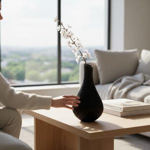 Puede incluir: Un jarrón negro texturizado con cuello delgado y cuerpo redondeado, que sostiene una rama de flores blancas. El jarrón está sobre una mesa de centro de madera clara. La escena está ambientada en un salón moderno con una gran ventana.