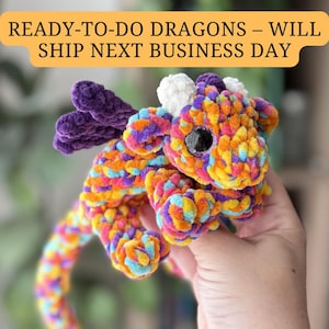 May include: A colorful, handmade crochet dragon with a vibrant mix of orange, yellow, purple, and blue yarn. The dragon has large, glittery eyes and a whimsical design. The text "READY-TO-DO DRAGONS - WILL SHIP NEXT BUSINESS DAY" is in the image.