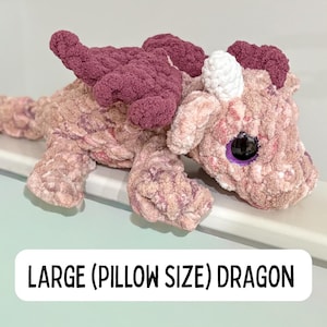 May include: A large, plush dragon in shades of pink and mauve, with a white horn and large black eye with a purple iris. The dragon has textured wings and limbs. Text on the image reads "LARGE (PILLOW SIZE) DRAGON."