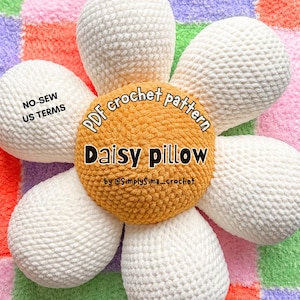 May include: A white crochet daisy pillow with a yellow center. The pillow is on a colorful checkered background. The text "PDF crochet pattern Daisy pillow by @SimplySima_crochet" is on the pillow.