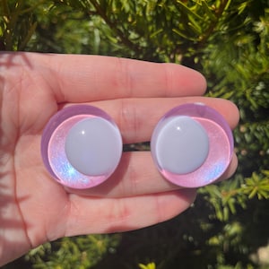 May include: Two round, iridescent purple and white plugs. The plugs have a white center and a purple outer ring with a shimmering, iridescent effect. The background is a blurred view of green foliage.