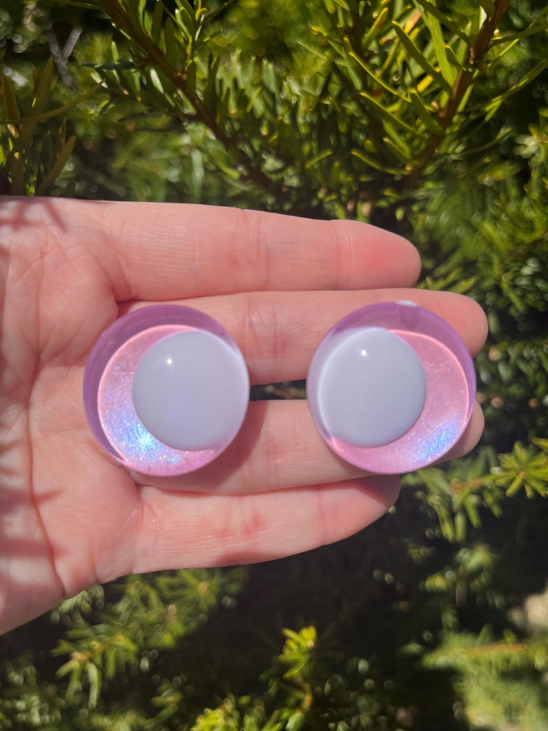 May include: Two round, iridescent purple and white plugs. The plugs have a white center and a purple outer ring with a shimmering, iridescent effect. The background is a blurred view of green foliage.
