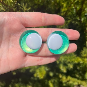 May include: Two round, green and white earrings. The earrings have a white center and a green, iridescent outer ring. The earrings are held in a hand against a blurred green background.