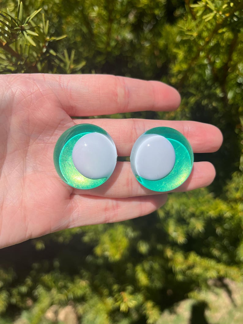 May include: Two round, green and white earrings. The earrings have a white center and a green, iridescent outer ring. The earrings are held in a hand against a blurred green background.