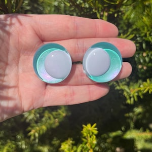 May include: Two round, iridescent teal and white earrings. Each earring features a white, circular center surrounded by a teal ring with a shimmering, iridescent effect. The earrings are set against a backdrop of green foliage.