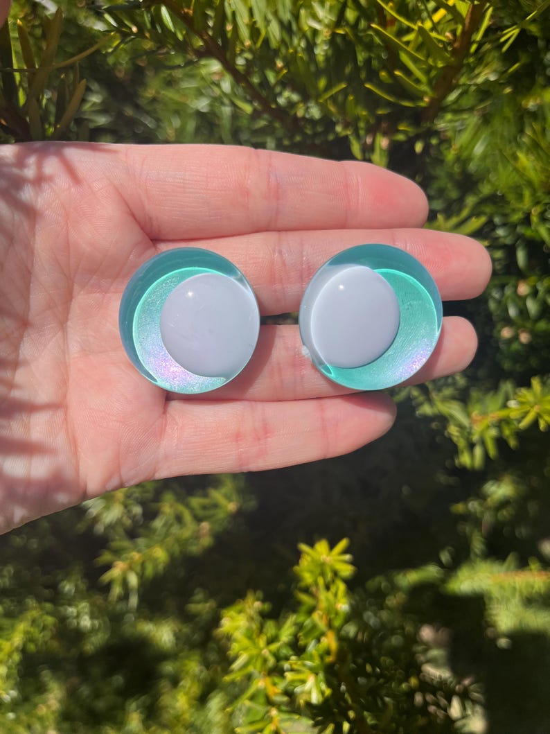 May include: Two round, iridescent teal and white earrings. Each earring features a white, circular center surrounded by a teal ring with a shimmering, iridescent effect. The earrings are set against a backdrop of green foliage.