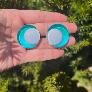 May include: A pair of round, teal and white earrings. Each earring features a white center surrounded by a teal ring and a clear outer layer. The earrings are held in a hand against a backdrop of green foliage.