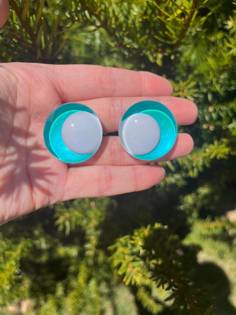 May include: A pair of round, teal and white earrings. Each earring features a white center surrounded by a teal ring and a clear outer layer. The earrings are held in a hand against a backdrop of green foliage.