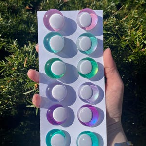 May include: A collection of twelve colorful, round, iridescent cabochons with a white center, arranged on a white card. The cabochons feature a variety of colors, including pink, teal, green, purple, and blue.