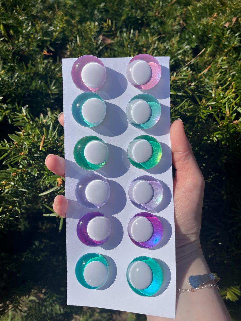 May include: A collection of twelve colorful, round, iridescent cabochons with a white center, arranged on a white card. The cabochons feature a variety of colors, including pink, teal, green, purple, and blue.