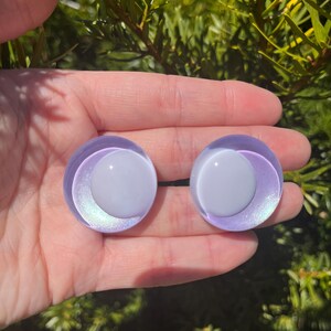 May include: Two round, iridescent purple and white glass eye cabochons. The eyes have a white center and a purple outer ring with a subtle, shimmering effect. The cabochons are held in a hand against a backdrop of green foliage.