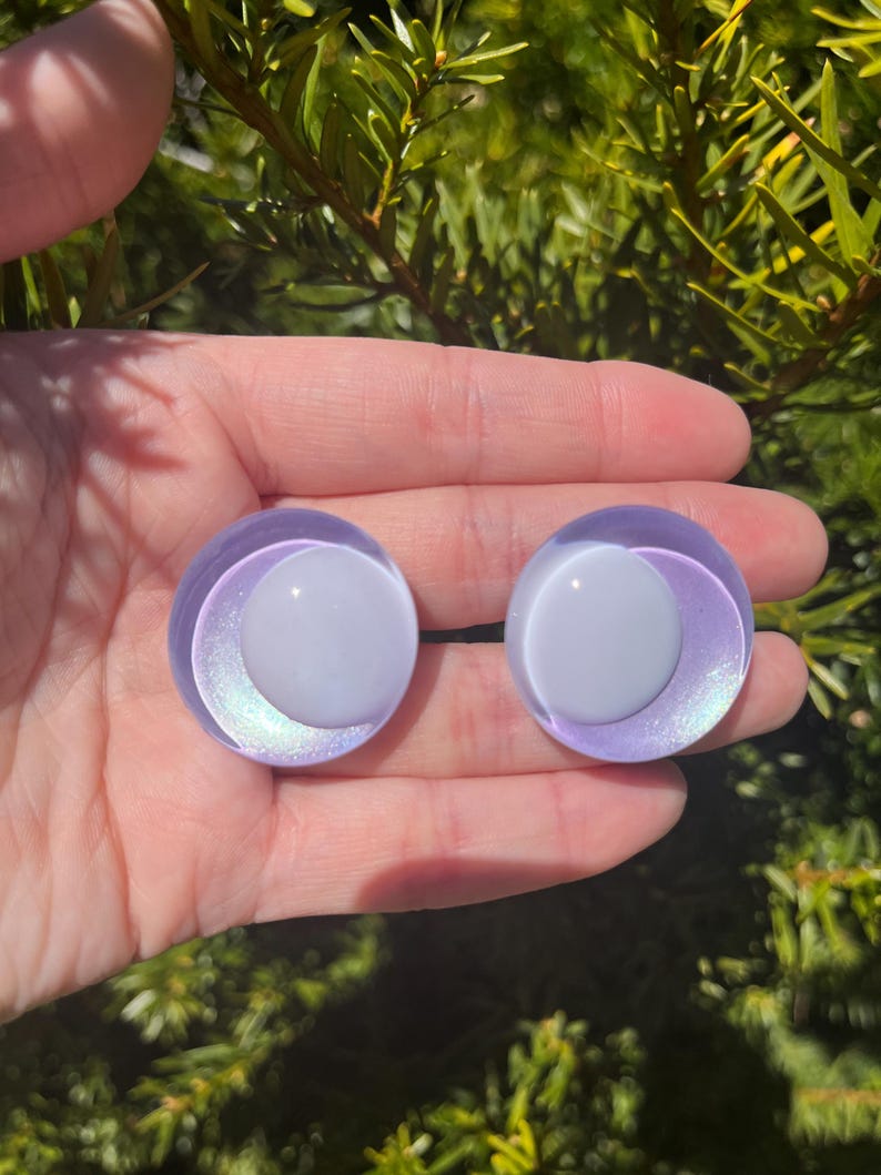 May include: Two round, iridescent purple and white glass eye cabochons. The eyes have a white center and a purple outer ring with a subtle, shimmering effect. The cabochons are held in a hand against a backdrop of green foliage.
