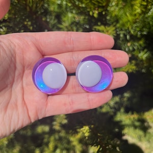 May include: A pair of iridescent purple and white ear gauges. Each gauge features a white center surrounded by a gradient of purple and blue. The gauges are round and appear to be made of glass or a similar material.