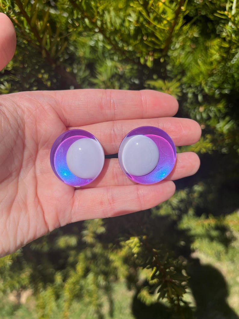 May include: A pair of iridescent purple and white ear gauges. Each gauge features a white center surrounded by a gradient of purple and blue. The gauges are round and appear to be made of glass or a similar material.