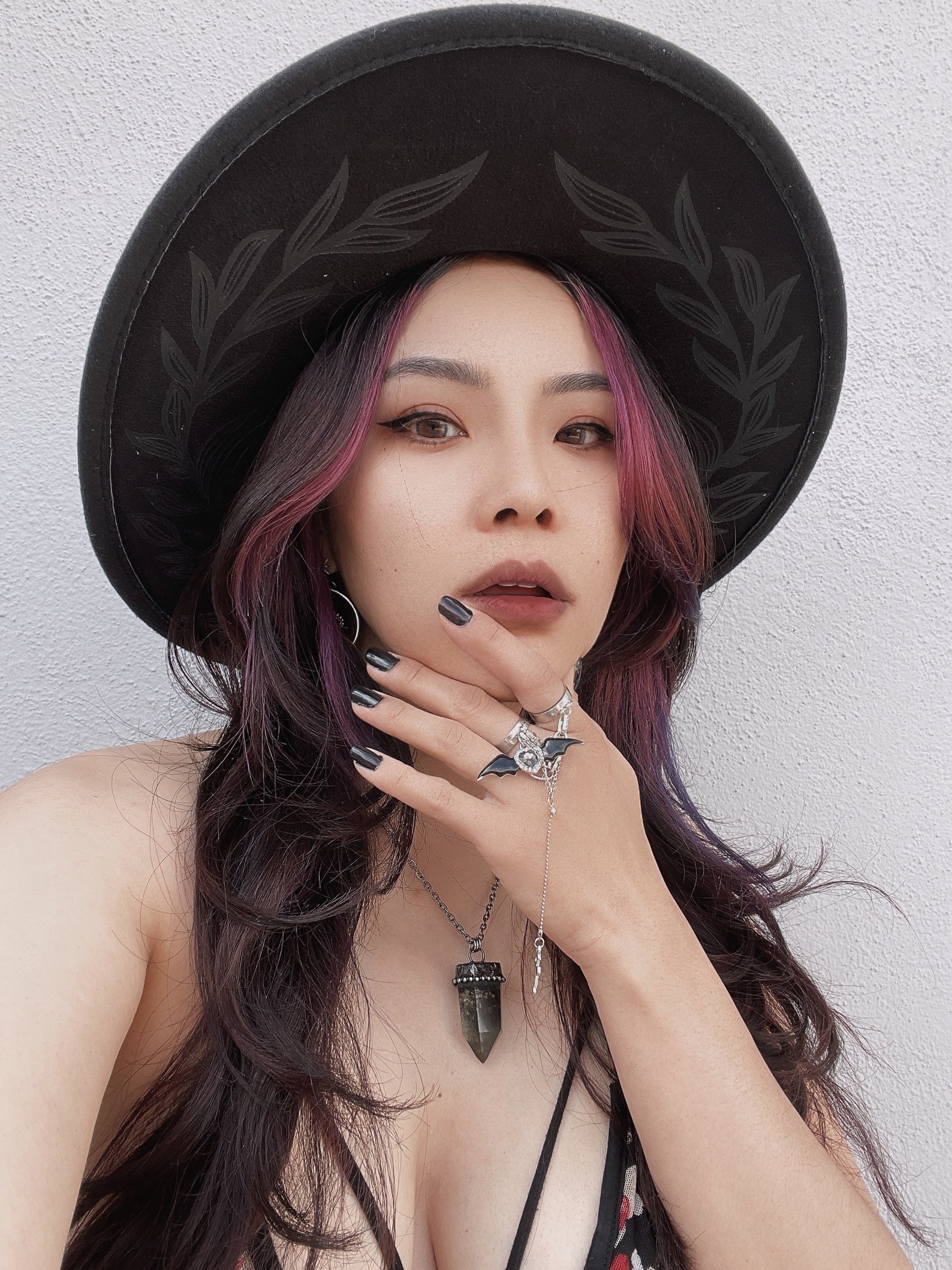 Goth woman in etched wide-brim hat with bat jewelry and crystal pendant, serving fierce summer goth style with mystical flair.