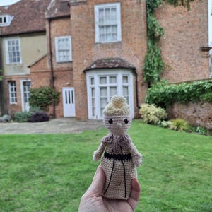 May include: A small, handmade crochet doll in a light beige dress, with a pink and black shawl and a light blonde bun. The doll is held in front of a brick building with a green lawn.