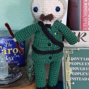 May include: A handmade crochet doll of a man with a gray beard and mustache, wearing a green suit with black accents. The doll holds two brown sticks. The background includes books and a sign that reads "DON'T LAUGH, IT'LL".