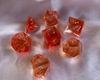 May include: Set of seven orange dice with white numbers. The dice are translucent and have a cloudy appearance. The dice are arranged in a circle on a white surface.
