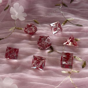 Op de afbeelding: Een set van zeven transparante dobbelstenen met rode nummers op een roze tule achtergrond. De dobbelstenen zijn in verschillende vormen en maten, waaronder een d4, d6, d8, d10, d12 en d20.