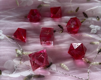 May include: A set of six pink dice with black numbers. The dice are on a pink and white fabric background.