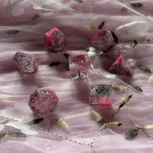 May include: A set of seven pink and clear dice with silver numbers. The dice are on a pink and white fabric background.