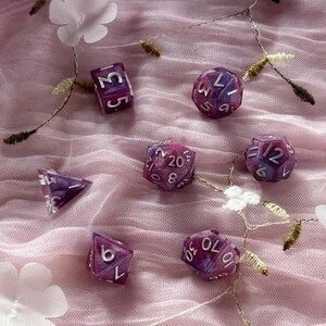Könnte beinhalten: Ein Satz von sieben lila und weißen Würfeln mit silbernen Zahlen. Die Würfel liegen auf einem rosa und weißen Tüllstoff.