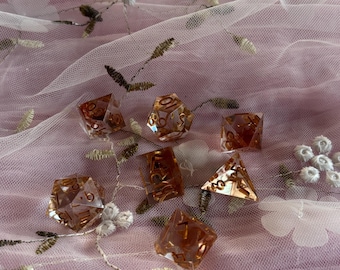 May include: A set of six clear, amber-coloured dice with gold numbers on a pink and white floral fabric background.