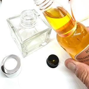 Könnte beinhalten: Eine klare Glasflasche mit schwarzem Verschluss und einem silbernen Ring. Die Flasche ist mit einer gelben Flüssigkeit gefüllt und trägt ein Etikett mit der Aufschrift "Zimt-Diffusor".