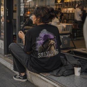 Op de afbeelding: Een zwart t-shirt met een afbeelding op de achterkant van een slak met een paarse muts. De tekst "LIVE SLOW OR WHENEVER" staat boven de slak en het woord "SNAILD IT" is in grote, verweerde witte letters onder de slak gedrukt.