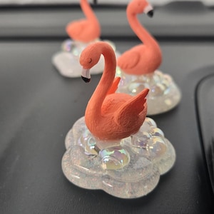 May include: Three coral-colored flamingo figurines with black beaks and white accents. Each flamingo is perched on a cloud-shaped base with iridescent glitter. The figurines are displayed on a dark surface.