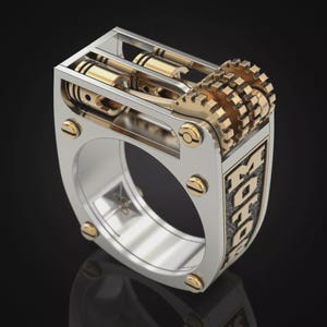 Puede incluir: Un anillo de tono plateado con un diseño de motor mecánico que presenta pistones y engranajes dorados en la parte superior. El lateral del anillo muestra la palabra MOTORS en letras doradas en relieve sobre un fondo oscuro y texturizado.
