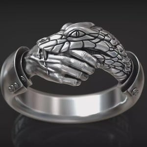 May include: A silver-toned ring featuring a detailed sculptural design of a human hand grasping the head of a snake. The snake has textured scales and a visible eye, while the hand appears to be emerging from a cuff-like sleeve detail on the band.