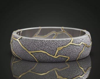 May include: A band ring featuring a textured, dark grey, granular surface finish contrasted by smooth, polished gold-colored lines that traverse the exterior in a pattern resembling cracks or fissures across the entire circumference of the band.