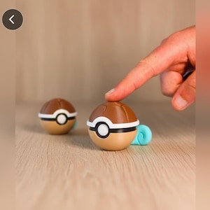 Puede incluir: Dos bolas novedosas marrones y beige con detalles blancos y una base azul claro, una de ellas tocada por un dedo. Las bolas tienen un diseño circular con una banda blanca y un centro negro. Las bolas están sobre una superficie marrón claro.