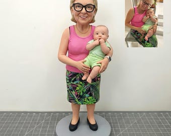 May include: A figurine of a woman wearing glasses, a pink tank top, and a skirt with a tropical leaf pattern, holding a baby in a green outfit. The figurine is standing on a gray circular base.